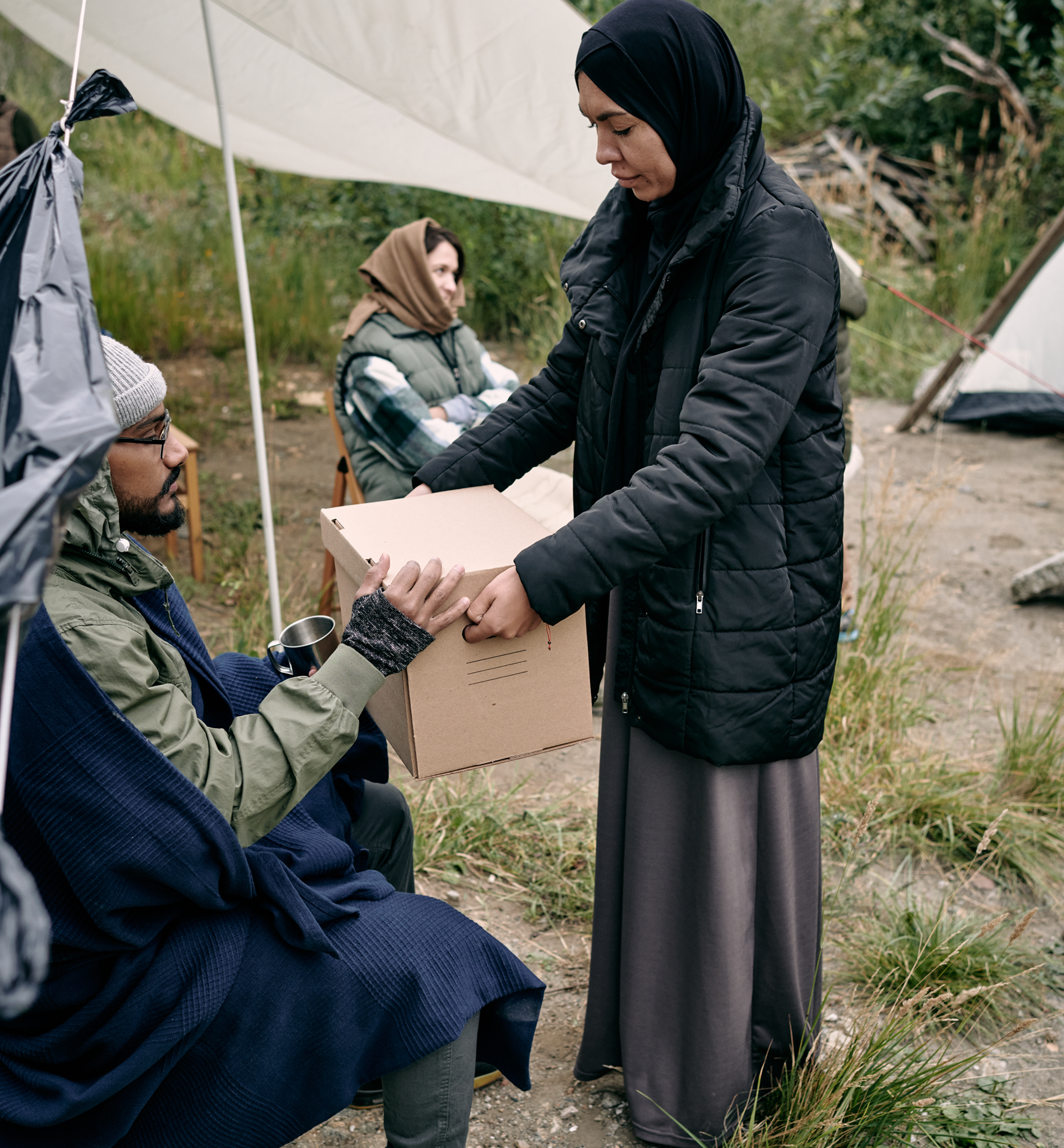 Woman handing box to seated man outdoors.